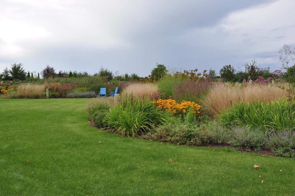 Ein liebevoll gestalteter Garten mit sattem, grünen Rasen, vielfältigen blühenden Stauden und Gräsern sowie zwei blauen Stühlen zwischen den Blumen - ein Beispiel für professionelle Gartenplanung und Pflanzplanung im Münsterland.