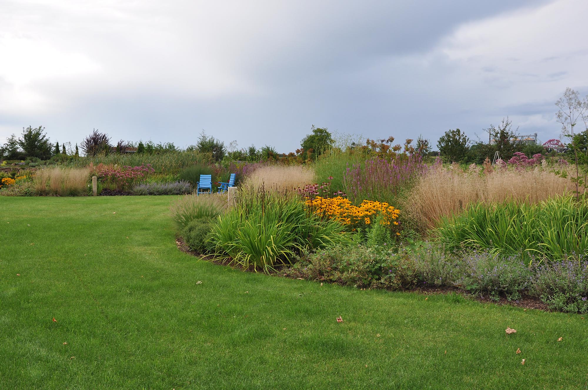 Ein liebevoll gestalteter Garten mit sattem, grünen Rasen, vielfältigen blühenden Stauden und Gräsern sowie zwei blauen Stühlen zwischen den Blumen - ein Beispiel für professionelle Gartenplanung und Pflanzplanung im Münsterland.