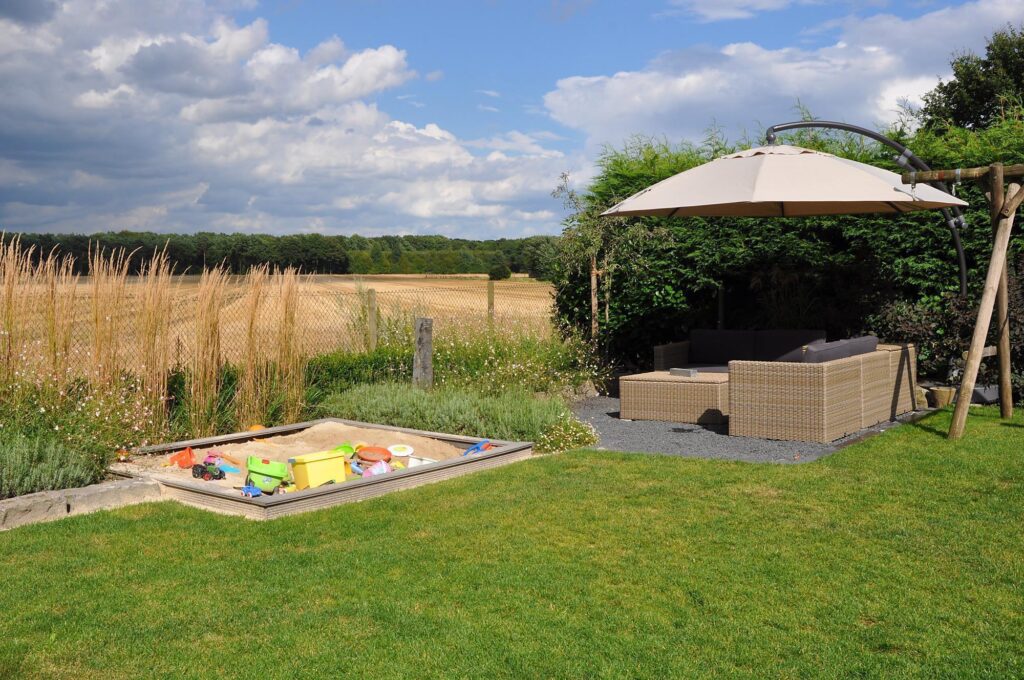 Ein liebevoll gestalteter Garten mit grünem Rasen, einem Sandkasten voller bunter Spielzeuge und einer gemütlichen Sitzgruppe unter einem Sonnenschirm - durch professionelle Gartenplanung und Pflanzplanung im Münsterland lebendig gestaltet.