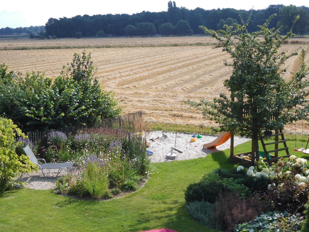 Ein familienfreundlicher Garten mit gepflegtem Rasen, gemütlichem Liegestuhl, harmonisch gestalteten Blumenbeeten sowie Spielbereich für Kinder mit Rutsche und Sandkasten - eingebettet in die Natur, ideal für professionelle Gartenplanung und Pflanzplanung im Münsterland.
