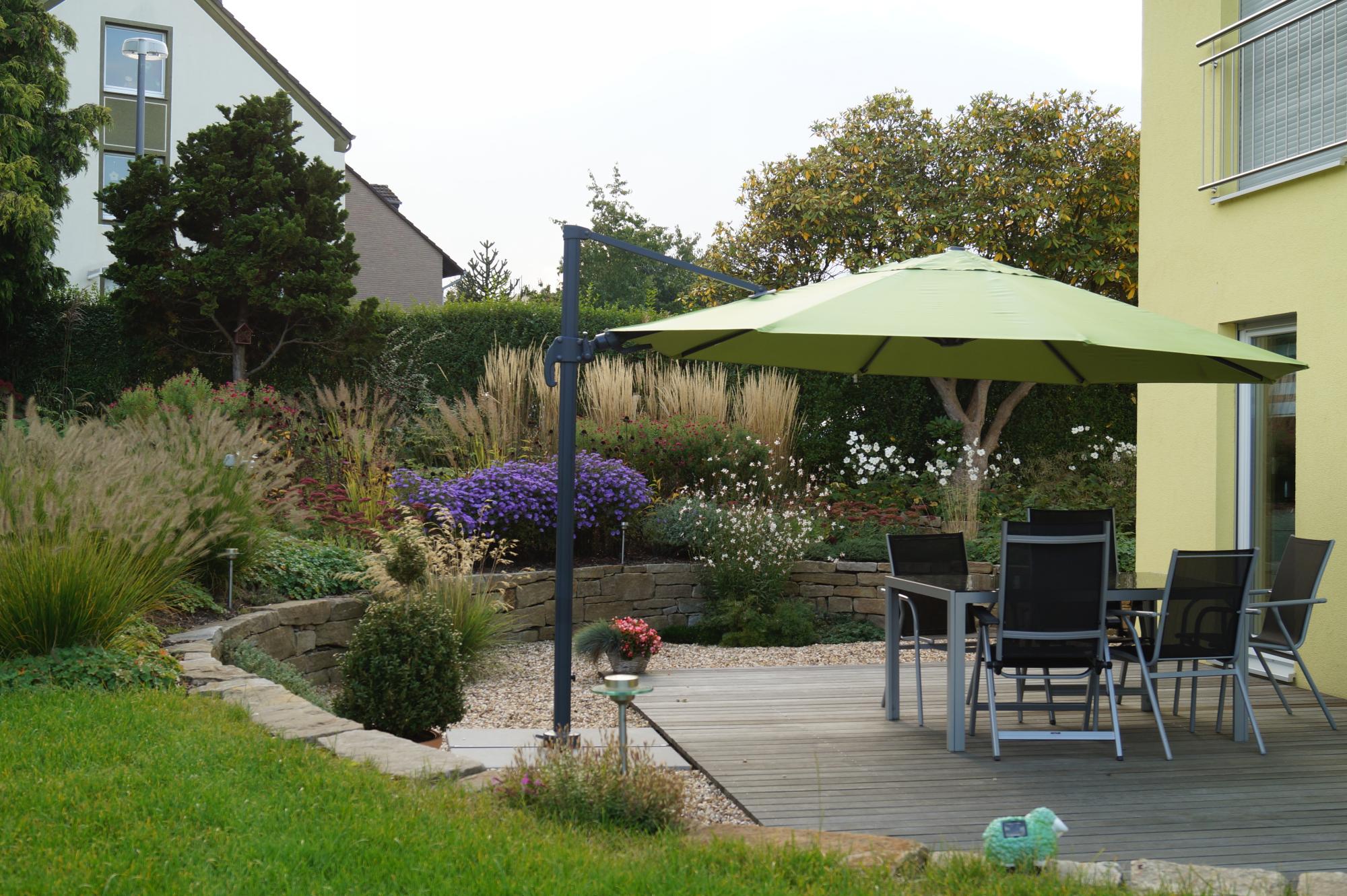 Auf einer stilvoll gestalteten Terrasse im Garten spendet ein großer grüner Sonnenschirm Schatten über einem Tisch mit vier Stühlen. Die professionelle Gartenplanung sorgt für strukturierte Begrünung mit Ziergräsern, Blühstauden und Natursteinmauer.