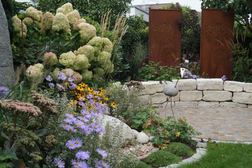 Ein liebevoll geplanter Garten im Münsterland mit blühenden Stauden, Ziergräsern und Sträuchern. Zwei hohe, rostfarbene Designelemente aus Metall, eine geschwungene Natursteinmauer, ein gepflasterter Bereich und eine Vogel-Skulptur sind zu sehen - Inspiration für professionelle Gartenplanung und individuelles Bepflanzungskonzept in Bocholt oder dem Kreis Borken.