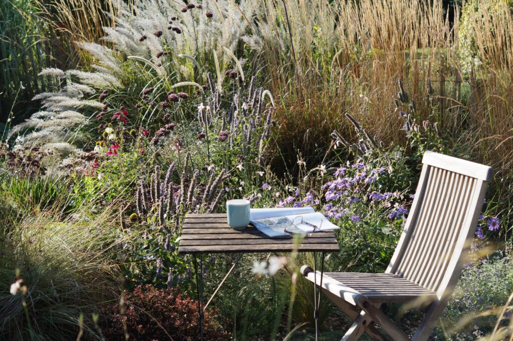 In einem naturnah gestalteten Garten, geplant durch professionelle Gartenplanung, stehen ein Holztisch und -stuhl zwischen hohen Gräsern und blühenden Wildstauden. Auf dem Tisch liegen Tasse, Brille und ein Buch - Inspiration für Ihre Pflanzplanung Garten.