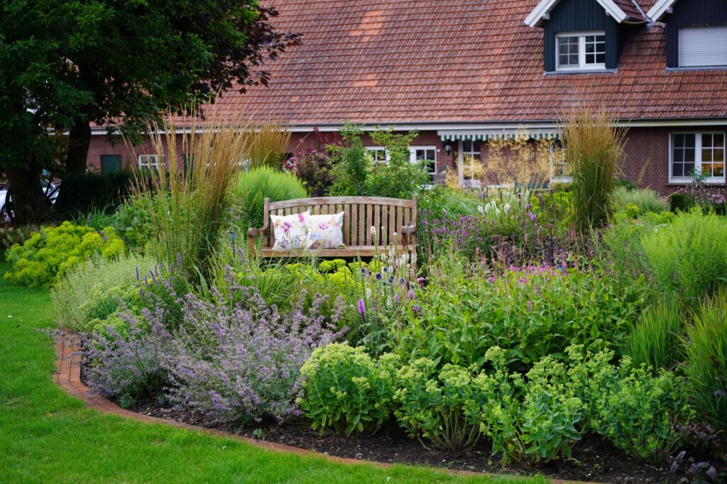 Eine gemütliche Holzbank mit floralen Kissen steht in einem liebevoll gestalteten Garten mit üppiger Bepflanzung und violetten Blüten. Dieser Garten vor einem Backsteinhaus zeigt die Wirkung professioneller Gartenplanung im Münsterland.