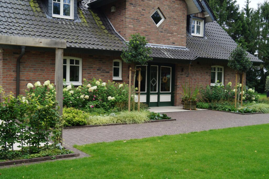 Ein Backsteinhaus mit dunklem Ziegeldach, weißen Sprossenfenstern und grün gerahmter Doppeltür. Im Vorgarten zeigen sich gepflegte Sträucher, Blumen, kleine Bäume und ein gepflasterter Weg - perfekt für professionelle Gartenplanung im Münsterland.