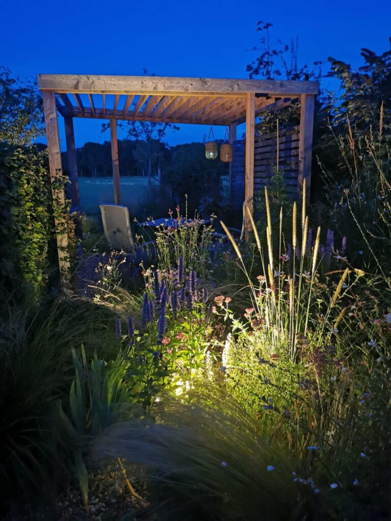 In einer professionell geplanten Gartenanlage im Münsterland steht eine stilvolle Holzpergola im abendlichen Licht. Sanfte Bodenstrahler inszenieren Ziergräser und Blühpflanzen - ein gelungenes Beispiel für moderne Garten- und Pflanzplanung.