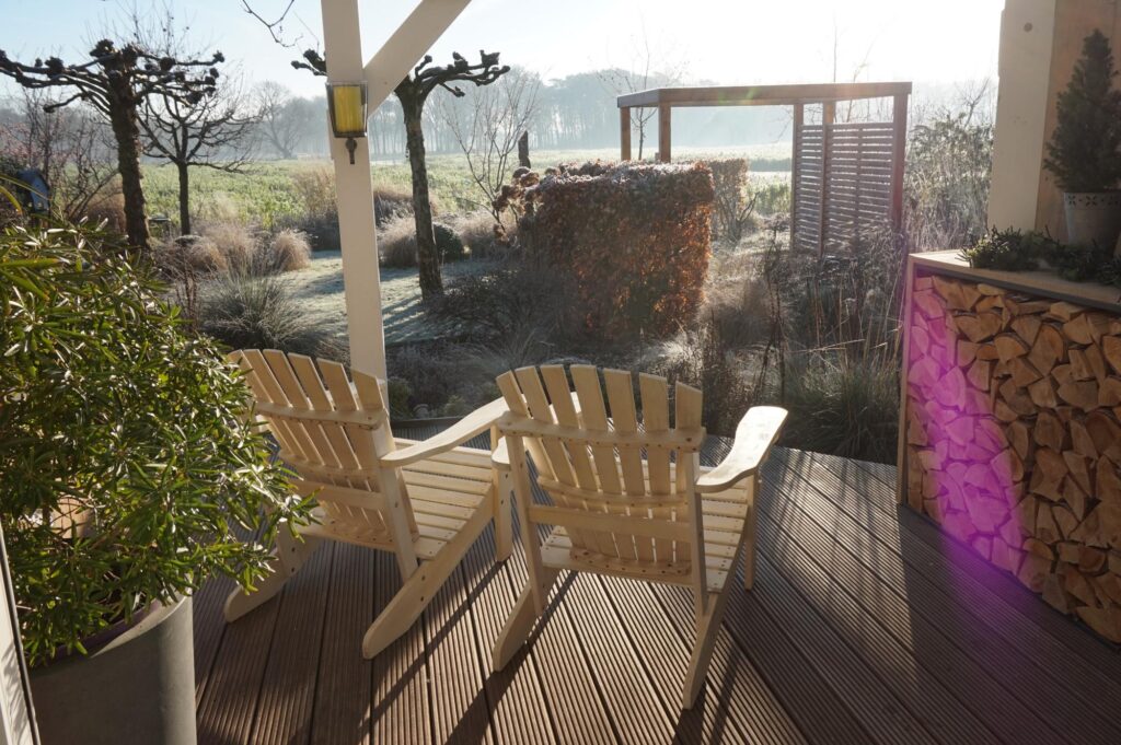 Zwei Holzstühle auf einer sonnigen Veranda blicken auf einen mit Raureif bedeckten Garten mit Bäumen, Sträuchern und gestapeltem Brennholz - ein stimmungsvolles Beispiel für professionelle Gartenplanung und Pflanzplanung im Münsterland.