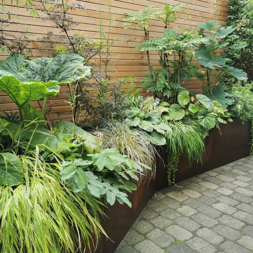 Ein stilvoller, moderner Garten mit üppig grünen Pflanzen in erhöhten Metall-Pflanzkübeln entlang eines Holzlattenzauns auf grauem Steinboden - ideal als Inspiration für professionelle Gartenplanung, Pflanzplanung oder ein Bepflanzungskonzept im Münsterland.