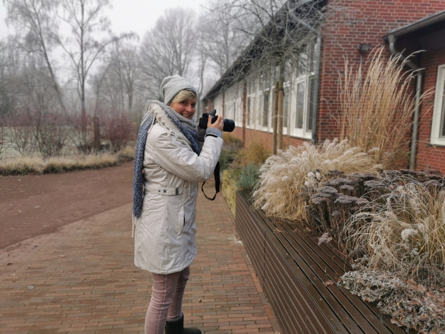 Eine Frau in Wintermantel, Schal und Mütze lächelt an einem frostigen Tag neben hohen Gräsern vor einem Backsteingebäude - ein inspirierender Moment für professionelle Gartenplanung, Pflanzplanung oder Bepflanzungskonzepte im Münsterland.