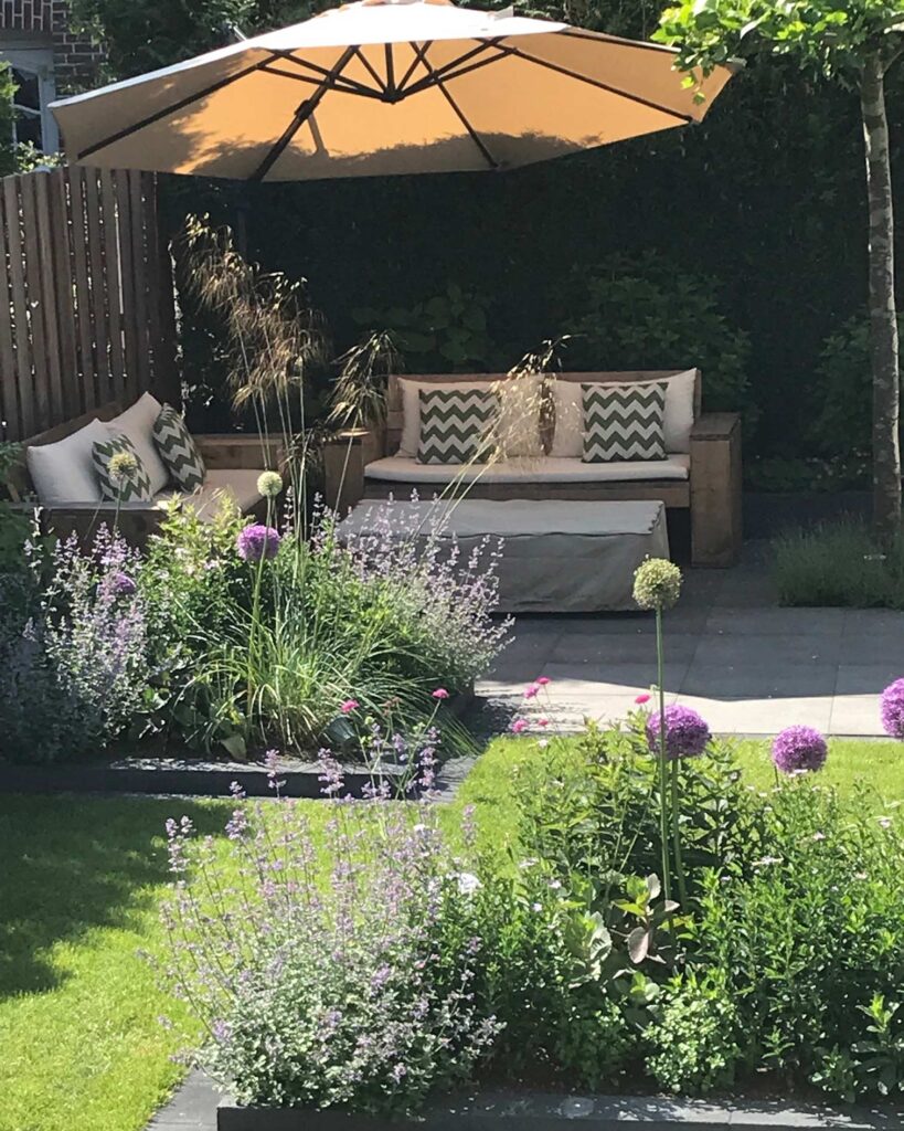 Eine einladende Garten-Terrasse mit beigem Outdoor-Sofa, Deko-Kissen und großem Sonnenschirm, umgeben von üppigen Pflanzen und blühenden lila Blumen - inspiriert von moderner Gartenplanung und individueller Pflanzplanung im Münsterland.