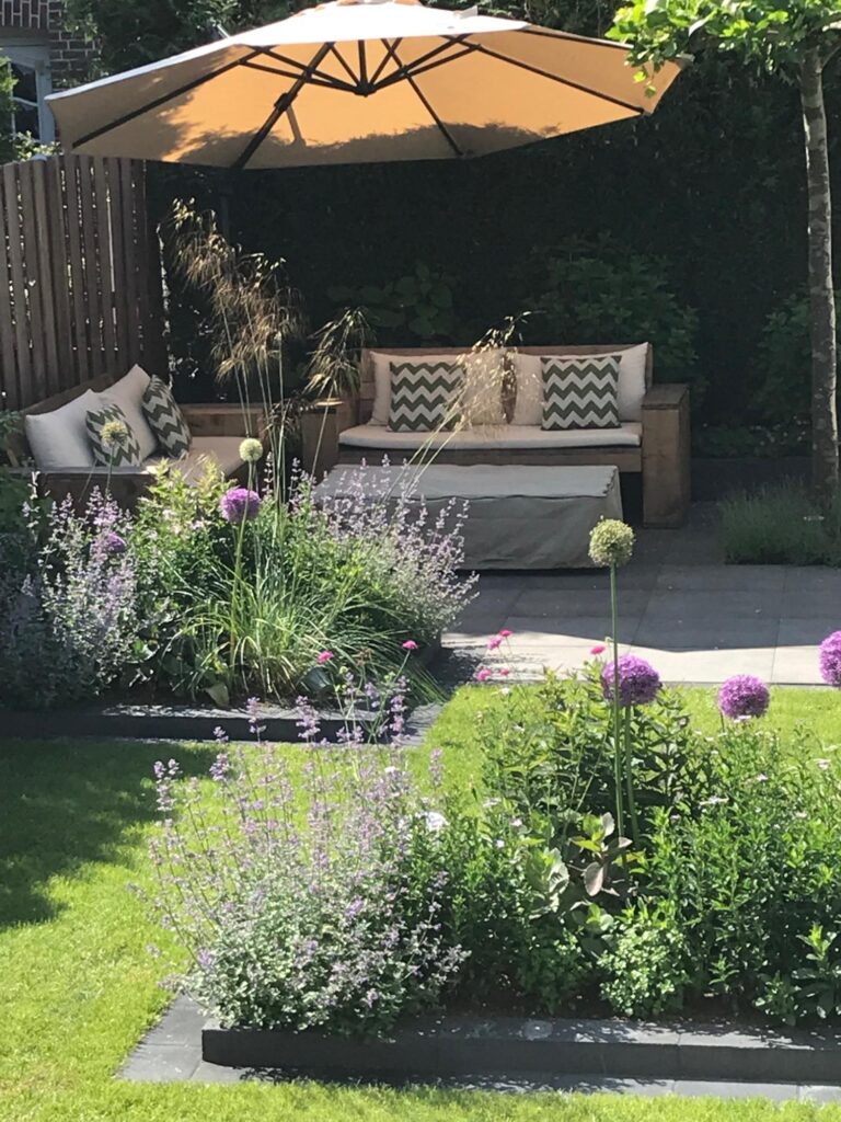 Eine gemütliche Sitzecke im Garten mit gepolsterter Bank und Kissen unter einem großen, hellen Sonnenschirm, umgeben von blühenden Pflanzen, gepflegtem Rasen und grünen Büschen - perfekt für Ihre professionelle Gartenplanung oder Pflanzplanung im Münsterland.