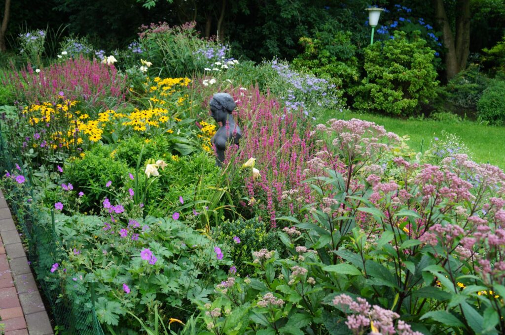 Ein liebevoll gestalteter Garten mit farbenfrohen Blumen in Gelb, Pink und Lila, grüner Bepflanzung und einer kleinen bronzenen Kinderstatue. Ein gepflasterter Weg führt entlang des Beetes - ein Beispiel für professionelle Gartenplanung im Münsterland.