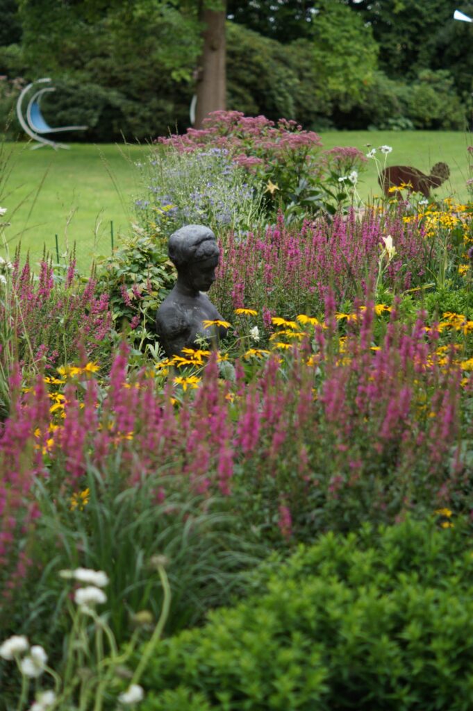 In einem sorgfältig gestalteten Garten mit professioneller Gartenplanung steht eine bronzene Büste zwischen hohen pinken und gelben Blüten. Umgeben von Bäumen und gepflegtem Rasen zeigt diese Szene, wie ein individueller Bepflanzungsplan den Garten neu erstrahlen lässt.