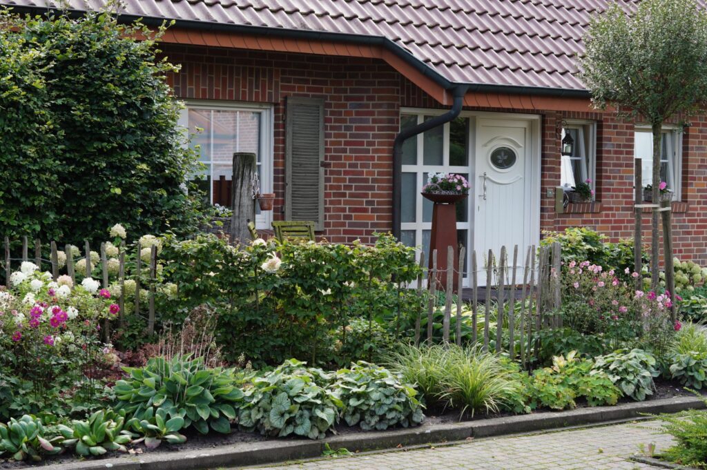 Ein Backsteinhaus mit weißer Tür steht hinter einem Holzpicketzaun und ist von einem farbenfrohen, blühenden Garten mit vielfältigen Pflanzen umgeben. Perfekt für professionelle Gartenplanung oder individuelle Bepflanzungskonzepte im Münsterland.