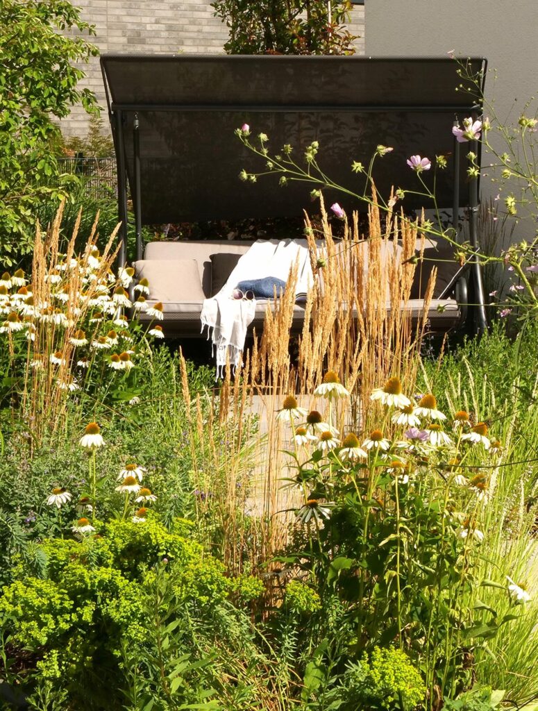Ein gemütliches Outdoor-Tagesbett mit Baldachin lädt zum Entspannen im sonnigen Garten ein, umgeben von hohen Gräsern, weißen Margeriten und üppiger Bepflanzung. So entsteht mit professioneller Gartenplanung und durchdachter Pflanzplanung ein Rückzugsort in Bocholt, im Kreis Borken oder im Münsterland. Ein weißes Kissen und eine Decke betonen das harmonische Bepflanzungskonzept - ideal für alle, die ihren Garten neu gestalten lassen möchten.