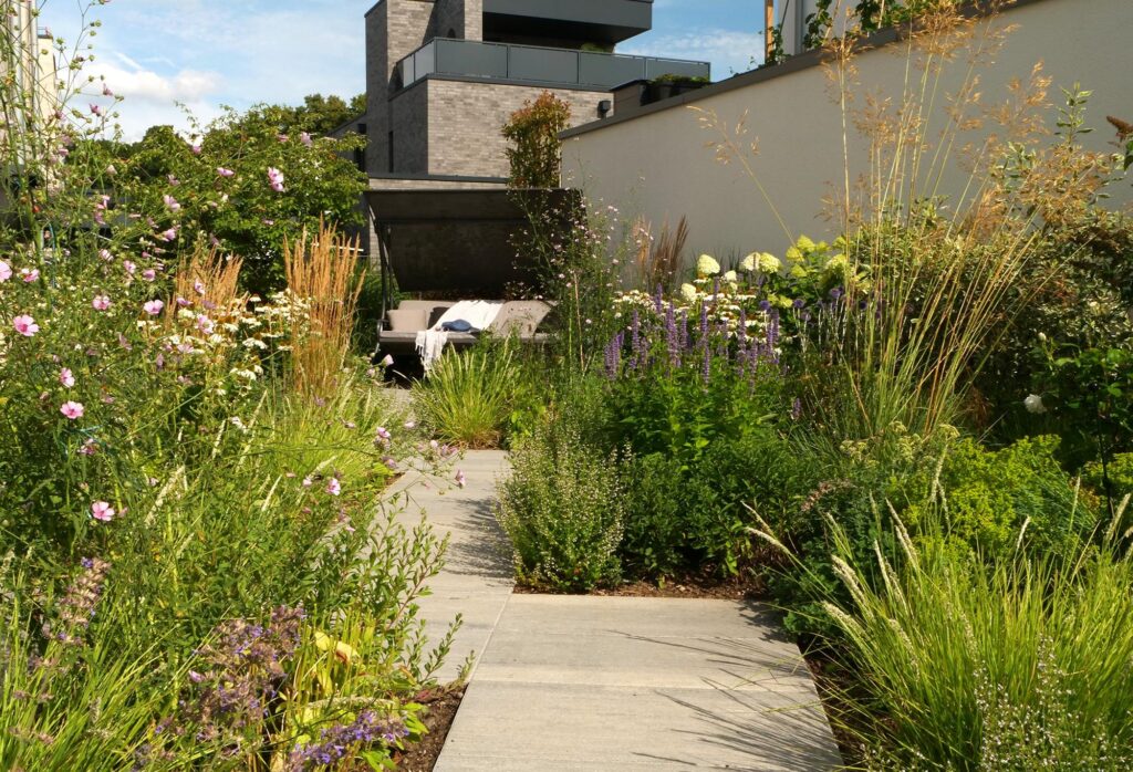 Professionell gestalteter Garten mit dekorativen Gräsern, Wildblumen und Sträuchern entlang eines gepflasterten Weges, der zu einer gemütlichen Sitzgruppe führt - inspirierendes Beispiel für individuelle Gartenplanung im Münsterland, Bocholt und Kreis Borken.